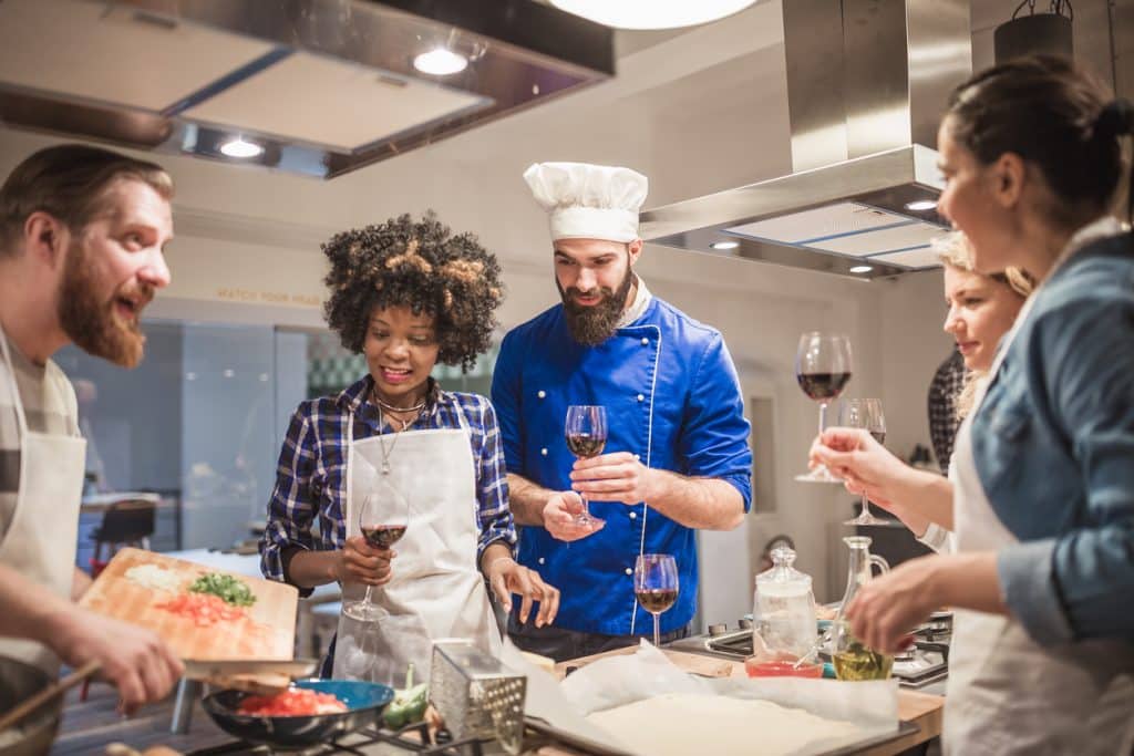 Diverse group of individuals gathers around a cooking station, having fun, cheering with red wine