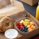 Open kraft breakfast box with everything bagel, fruit cup, cream cheese, and orange juice for a morning meeting