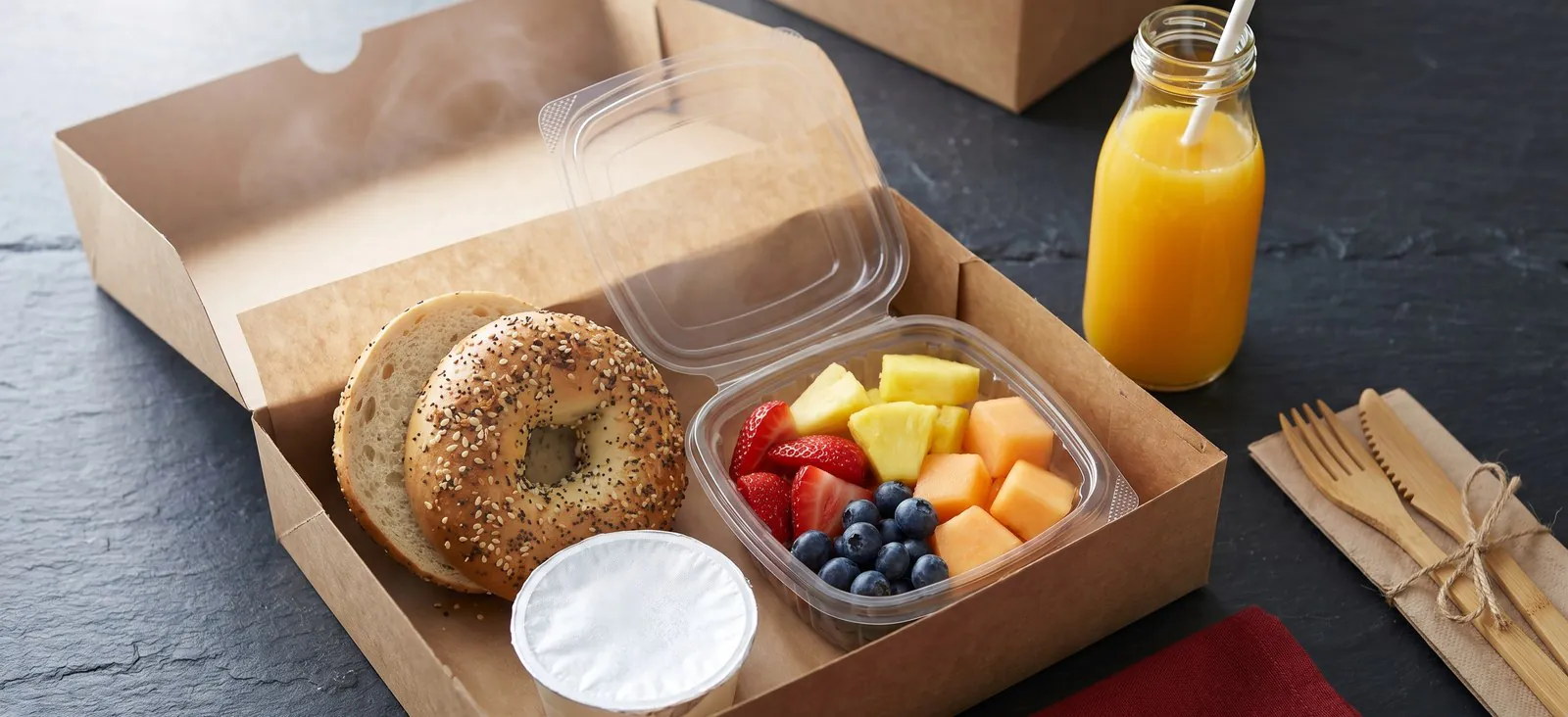 Open kraft breakfast box with everything bagel, fruit cup, cream cheese, and orange juice for a morning meeting