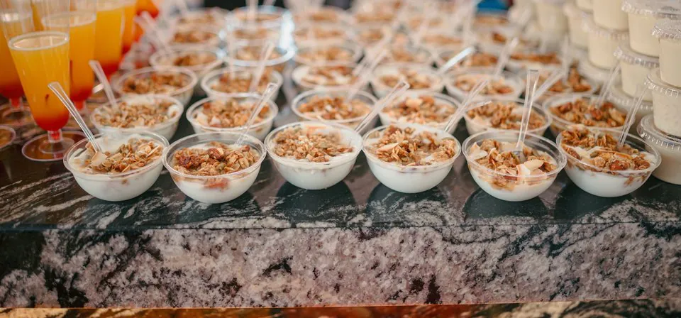 Rows of individual yogurt and granola parfait cups with juice cups on a marble counter for office breakfast catering