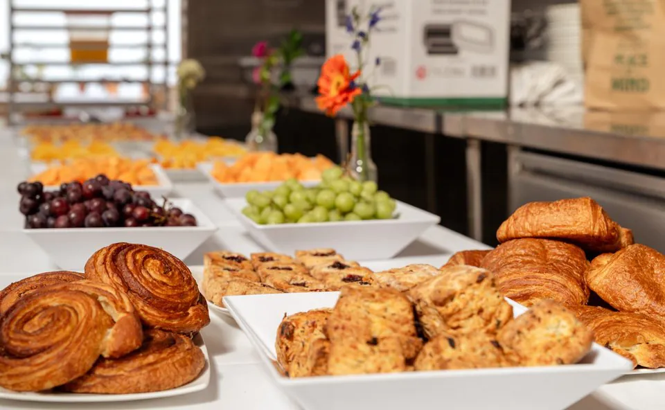 Continental breakfast catering spread with croissants, pastries, scones, fresh grapes, and melon on white square plates