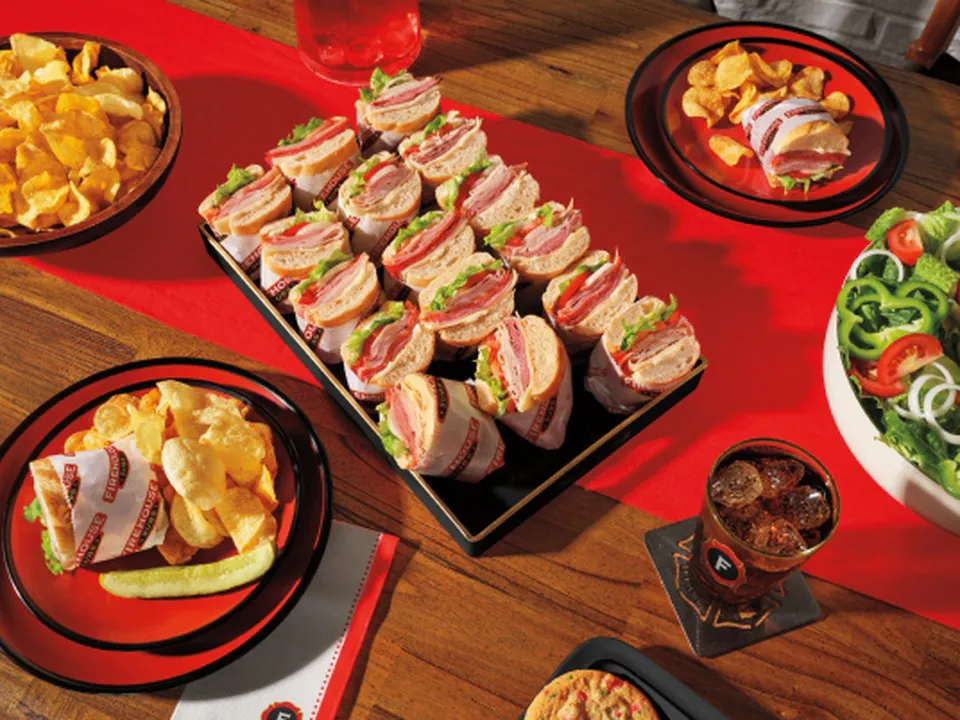 Sub platter of sliced sandwich halves arranged on a black serving tray with sides of chips and salad