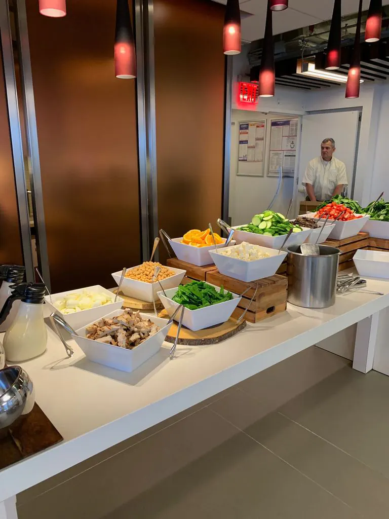 Office catering buffet with a naturally gluten-free spread: white bowls of chickpeas, oranges, fresh mozzarella, spinach, grilled chicken, cucumbers, peppers, with serving tongs ready