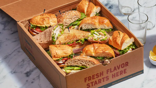 Open kraft catering box of assorted McAlister's Deli signature sandwiches sliced into halves on toasted bread, with empty pebbled glasses to the side