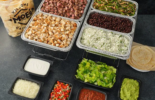 Moe's Southwest Grill Fajita Bar catering setup with grilled chicken, steak, fajita peppers and onions, black beans, cilantro-lime rice, sour cream, cheese, lettuce, guacamole, salsa and pico de gallo, with soft flour tortillas and a chip bag