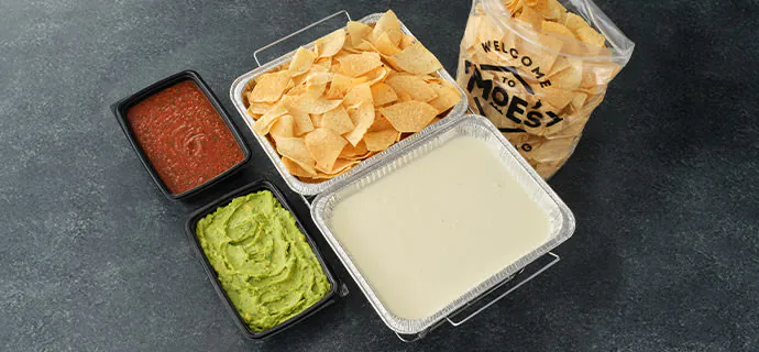 Moe's Famous Queso served in a foil catering pan alongside warm tortilla chips, red salsa and a side of guacamole