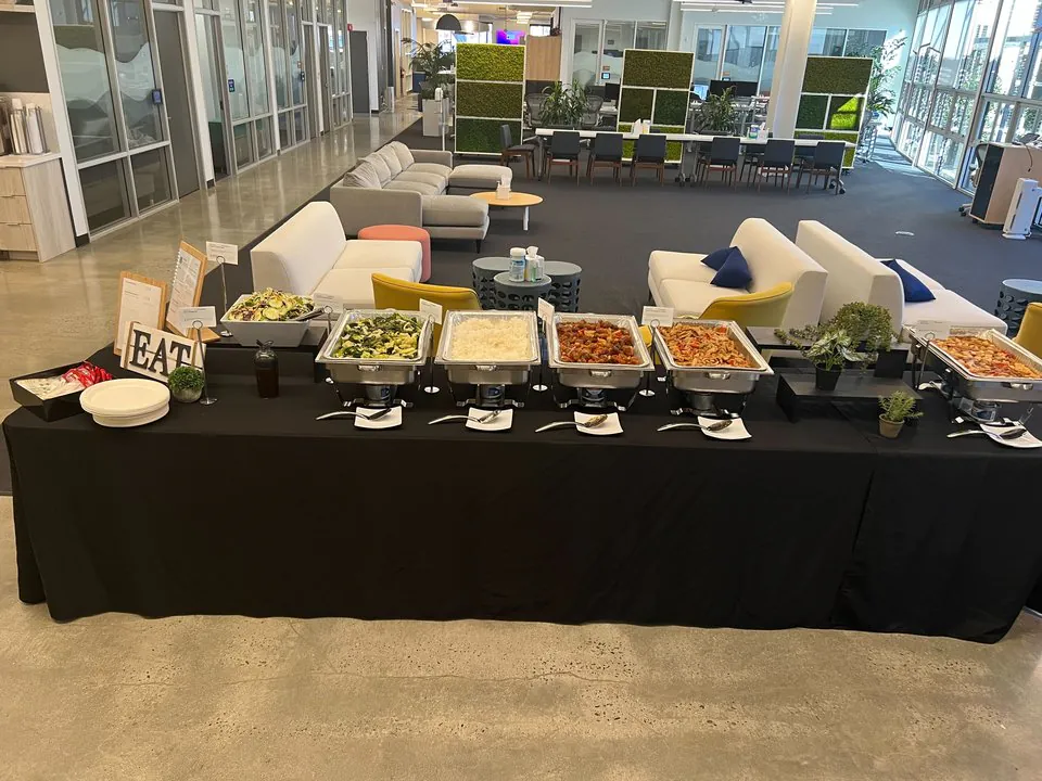 Office buffet line with Italian-style chafing trays of pasta, salad, and protein on a black-linen table in an open-plan workspace