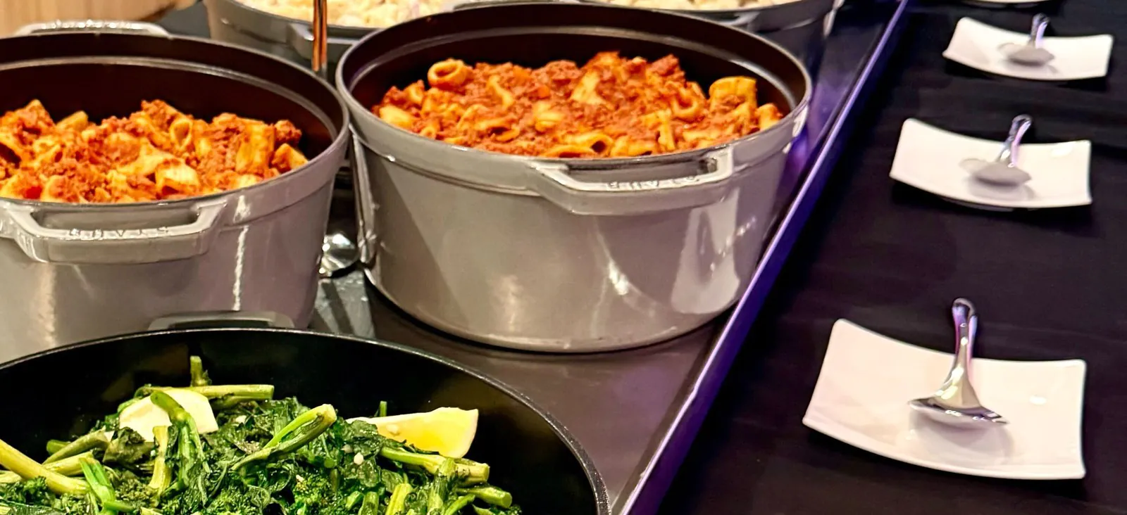 Italian-American catering spread with red-sauce rigatoni, baked ziti, and saut&eacute;ed broccoli rabe in serving pots on a black-linen buffet line
