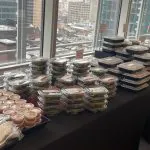 Stacked clear individually packaged boxed lunches with allergen placards and wrapped utensils on an executive office table with city view