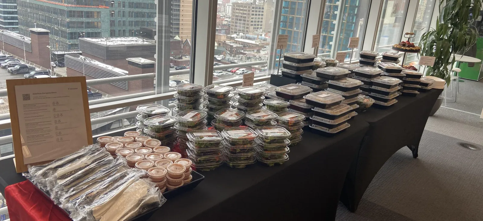 Stacked clear individually packaged boxed lunches with allergen placards and wrapped utensils on an executive office table with city view