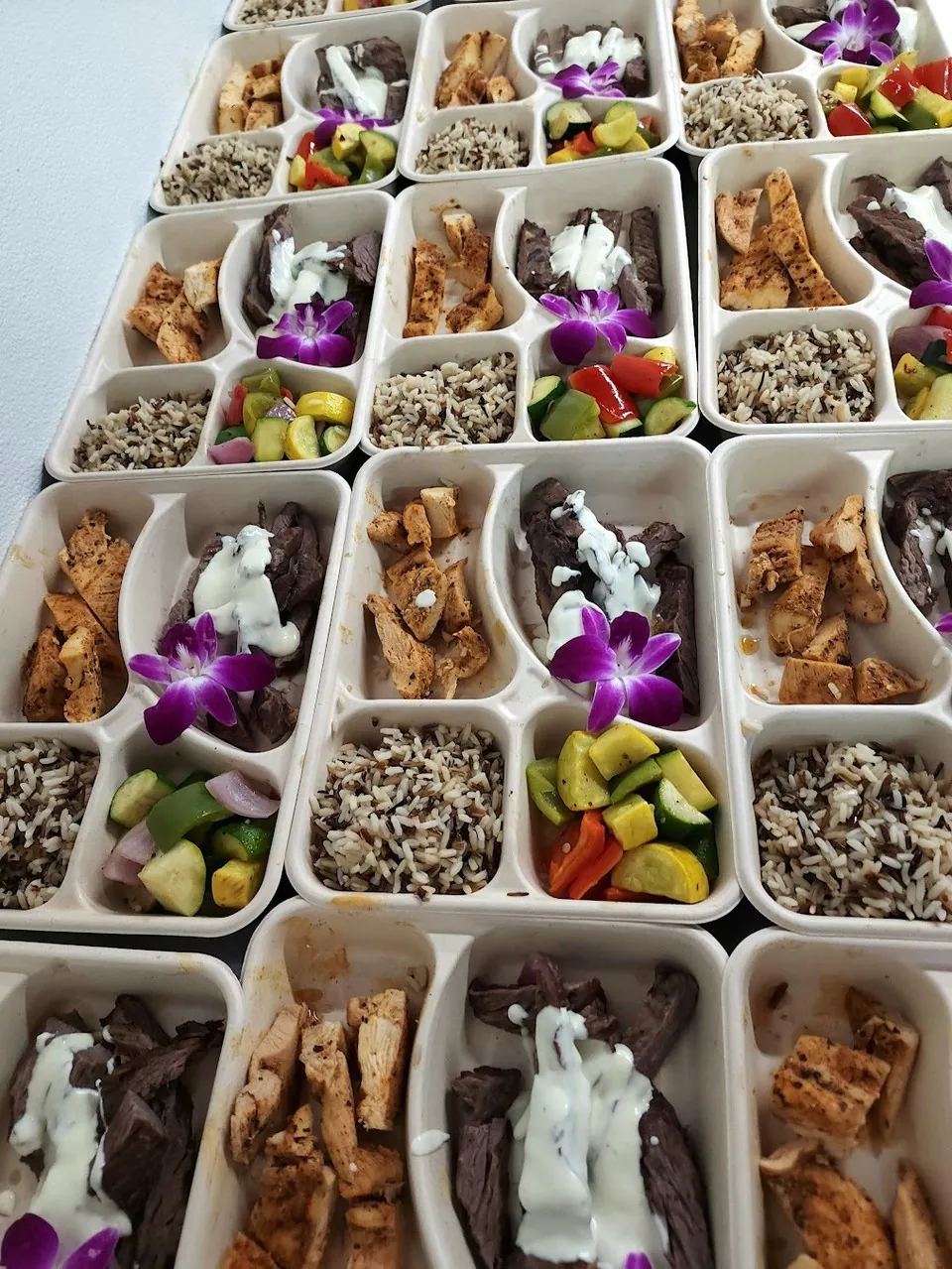 Overhead grid of compartmented bento boxes with grilled chicken, sliced steak, wild rice, and vegetables for office catering