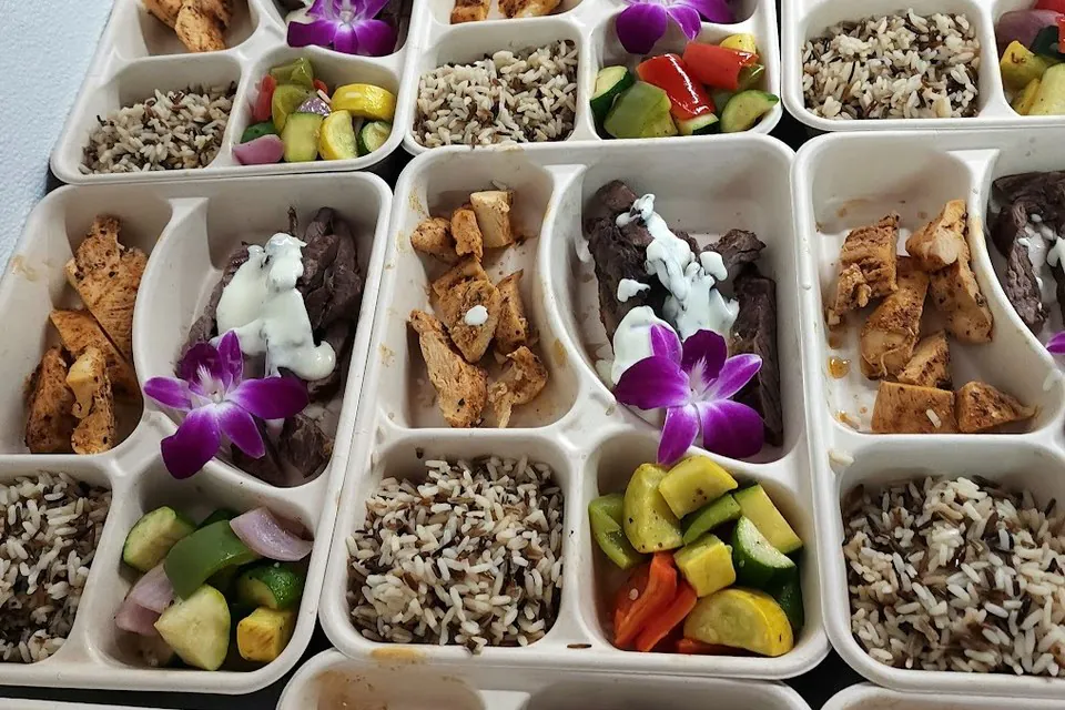 Overhead grid of compartment catering trays with sliced steak, grilled chicken, wild rice, and summer vegetables for an Atlanta office boxed lunch order