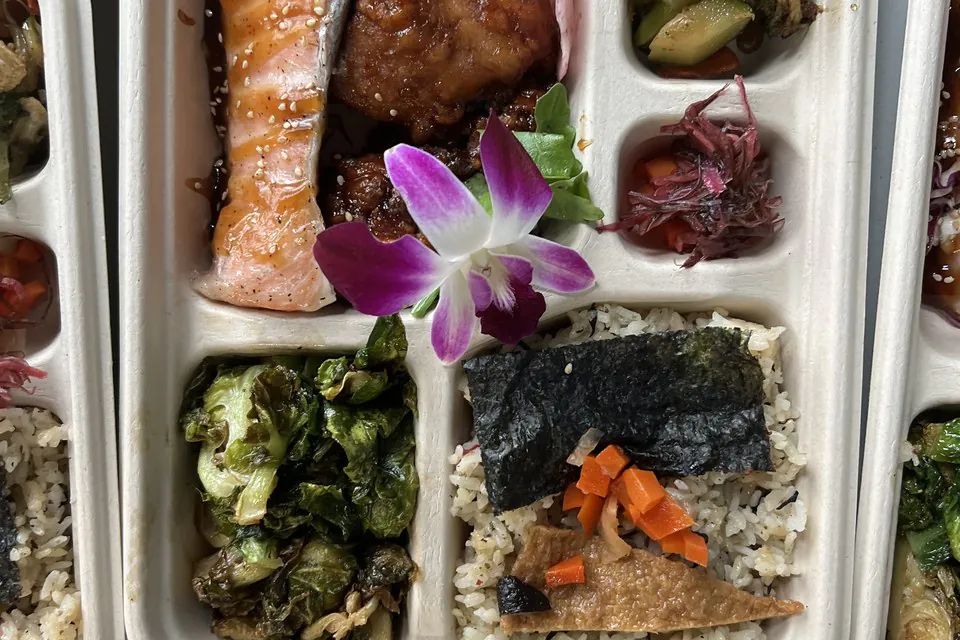 Overhead view of compartment bento boxes with salmon, fried chicken, brussels sprouts, and nori-topped rice for office boxed lunches in Austin
