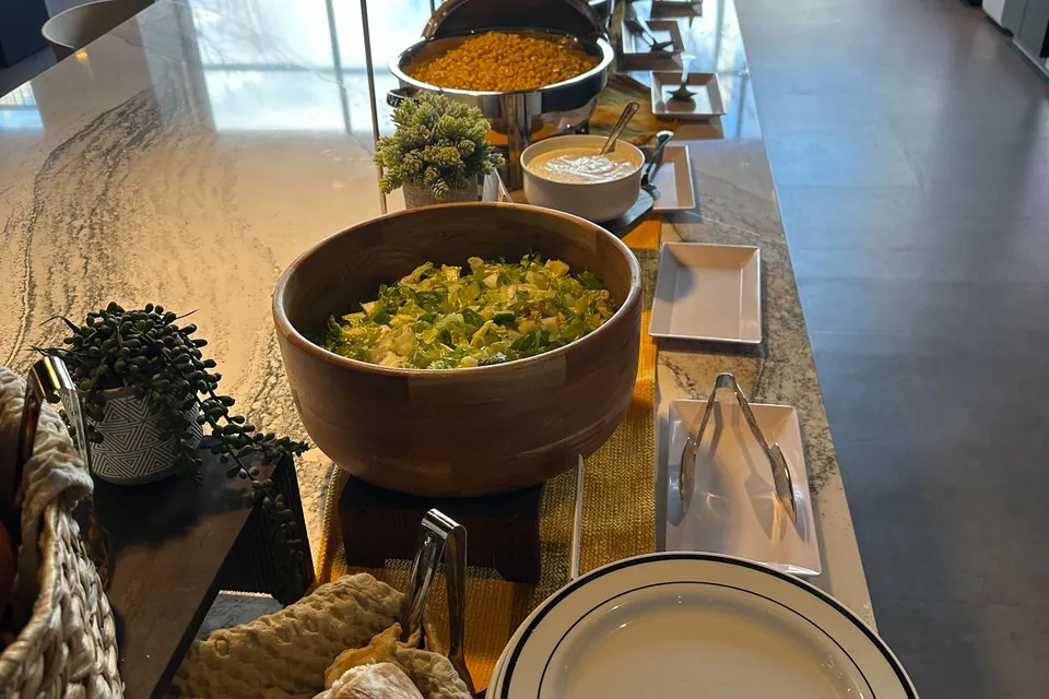 Office buffet line with wooden salad bowl, chafing dishes, plates, and bread basket on a marble counter