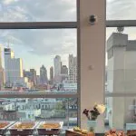 Real Zerocater office catering setup with chafing dishes, shared platters, fresh flowers, and an NYC skyline view through floor-to-ceiling windows. A representative ZC client offsite event.
