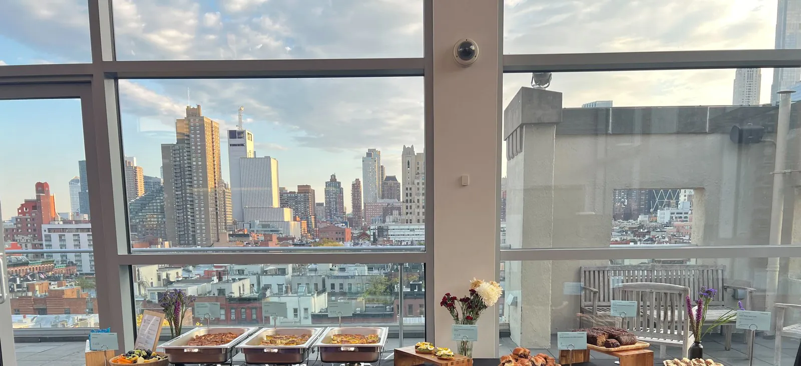 Real Zerocater office event catering setup: chafing dishes lined along a black-draped table, shared platters and fresh flowers, NYC skyline visible through floor-to-ceiling windows behind