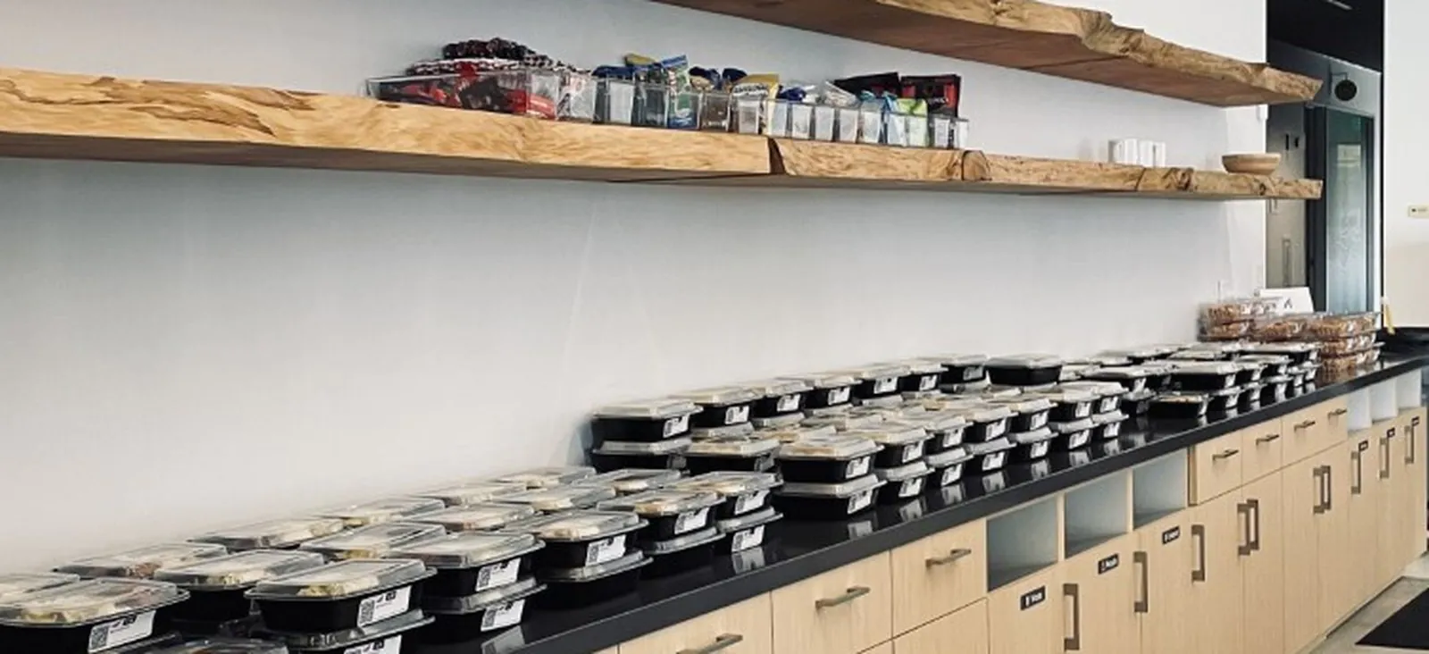 Real Zerocater office boxed lunch distribution at a national-chain scale Real Zerocater office boxed lunch setup: long black counter lined with rows of individually packaged catering meals under wood shelves with snacks, dessert tray on the right side