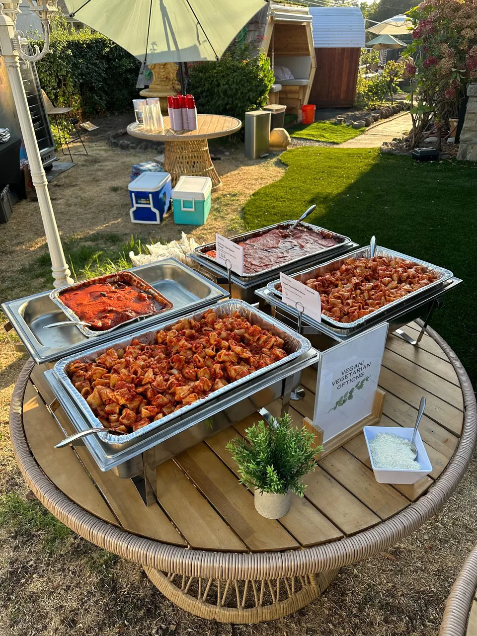 Overhead view of outdoor BBQ buffet chafers full of pulled pork, chicken, ribs and red sauces on a wood picnic table with cooler and patio umbrella in the background