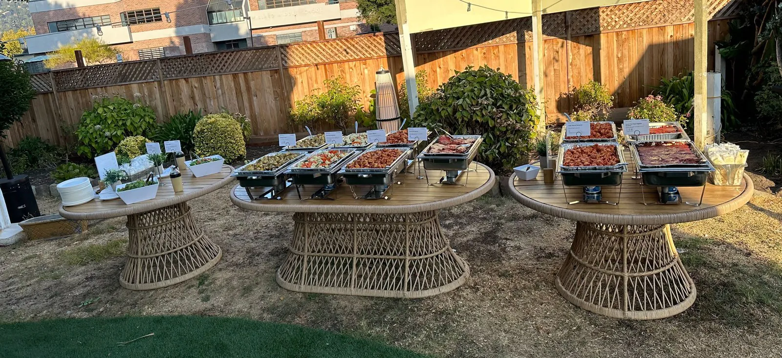 Outdoor company picnic catering buffet on three wicker tables in a backyard with chafers full of fresh fruit and proteins at golden hour