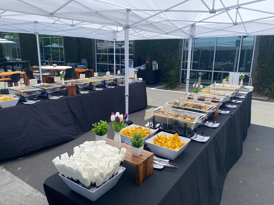 White rental tent over a long black-linen buffet table with multiple chafers, fresh tulip florals, and grab-and-go side stations for an outdoor company catering event