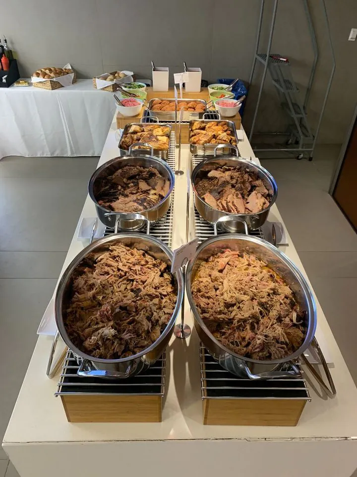Office BBQ catering buffet line with stainless chafing pans of pulled pork in the foreground, sliced brisket behind, and small sides of slaw and pickled red onions on a side table