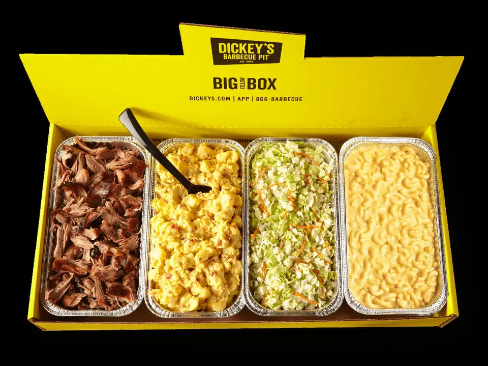 Top-down view of Dickey's Barbecue Pit Big Yellow Box catering package with four foil pans of pulled pork, potato salad, cole slaw, and mac and cheese in a yellow branded box