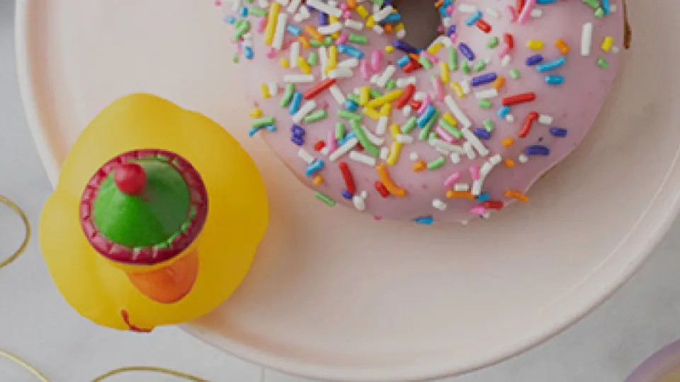 Pink-frosted rainbow-sprinkle Duck Donuts donut on a soft pink plate next to a yellow Ollie the Duck rubber duck mascot in a party hat, on a white marble counter