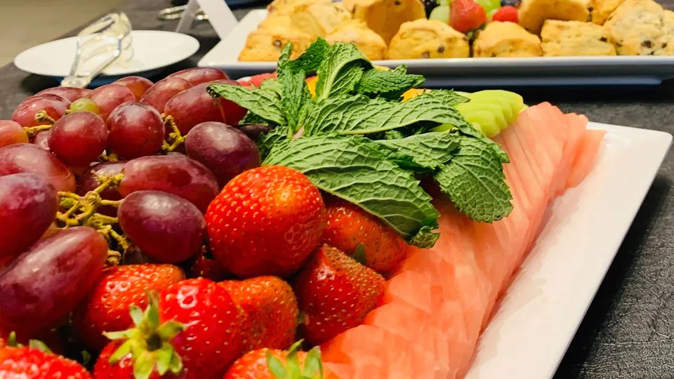 Catering platter of sliced Nova lox salmon with fresh mint, red grapes and strawberries, alongside a deli tray of pastries and scones in the background