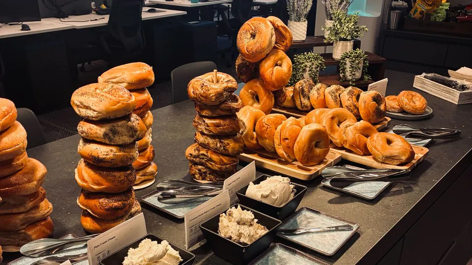 Office catering bagel and shmear bar with tall stacks of poppy seed, asiago, and plain bagels, a wooden bagel platter, and stoneware bowls of cream cheese on a dark stone counter