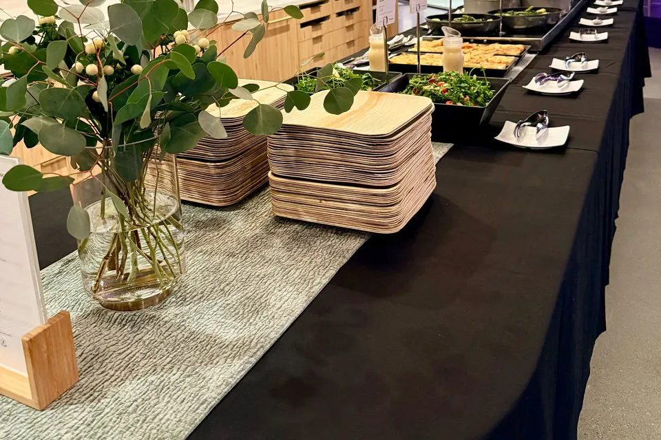 Buffet setup with stacks of wooden plates, eucalyptus centerpiece, and prep trays down a long black-draped office event table