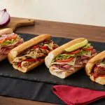 Mixed sub catering platter with hot and cold subs sliced into shareable portions on a black slate board, ramekins of chips and pickles, mustard, on a black-linen office buffet