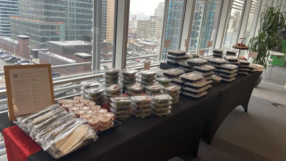 Stacks of individually packaged holiday lunches on a black-draped table beside a snowy city skyline view from a high-rise office