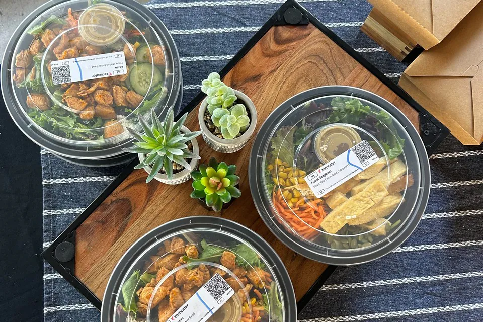 Overhead view of three sealed round salad bowls with chicken, mixed greens, corn, carrots, cucumber, and crispy strips on a wooden cutting board with succulents and kraft takeout boxes