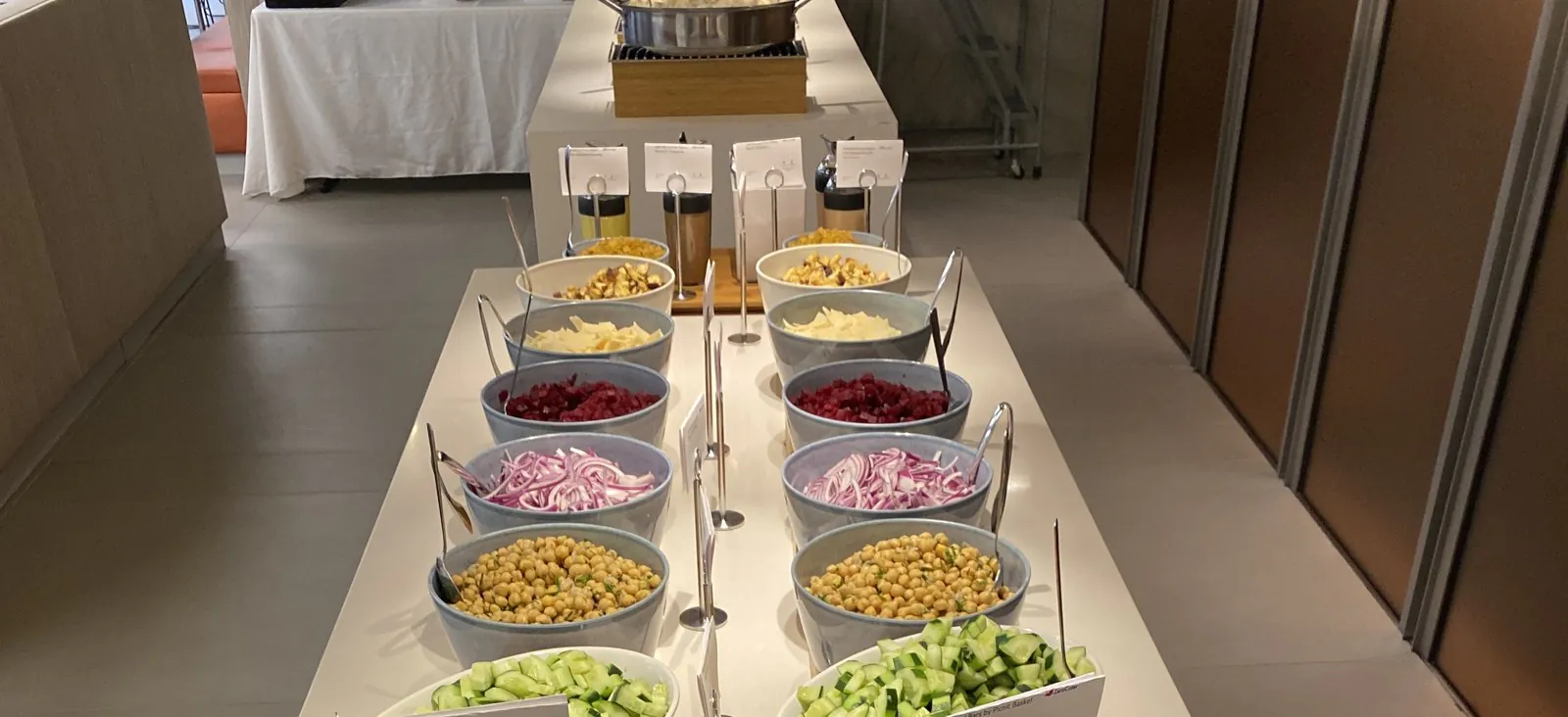 Office salad bar with white bowls of corn, cheddar, beets, red onions, chickpeas, cucumbers, and tomatoes on a white counter with stainless tongs