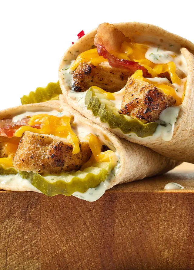 Two grilled chicken wraps with dill pickles, melted cheddar and creamy herb sauce on a wooden cutting board, Jason's Deli catering