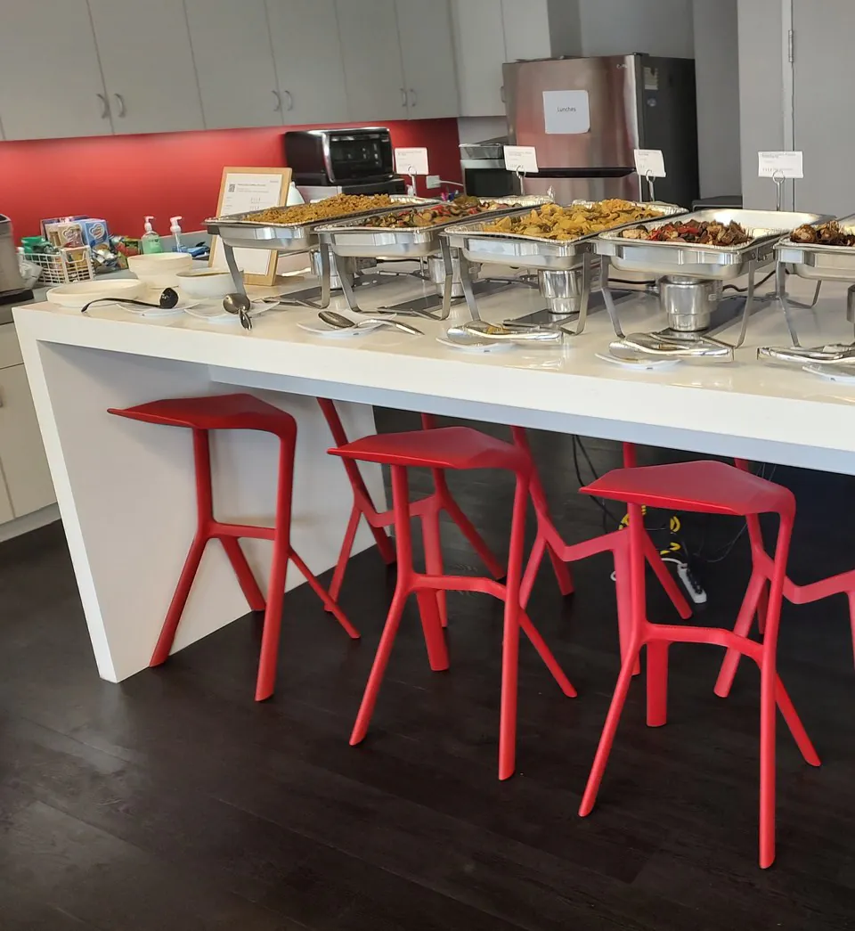 Office buffet line of chafing trays holding fried rice, chow mein and wok-tossed entrees on a white counter with red stools