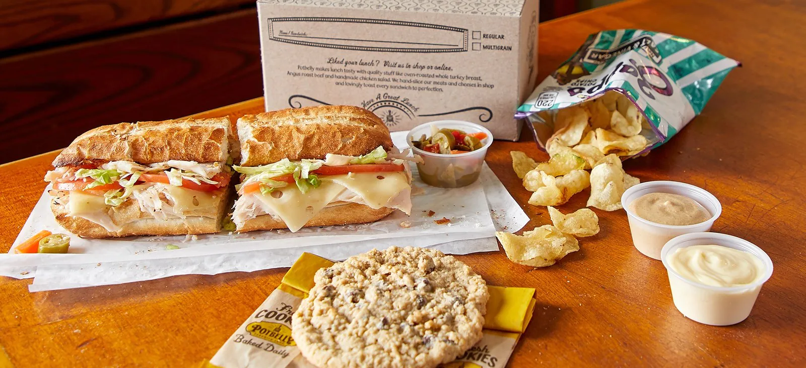 Open Potbelly Boxed Lunch on a wood table: signature halved warm-toasted long roll with turkey, ham, Swiss, lettuce, tomato and hot peppers on parchment, single-serve chips, Potbelly oatmeal chocolate-chip cookie, and ramekins of spreads next to the branded catering box