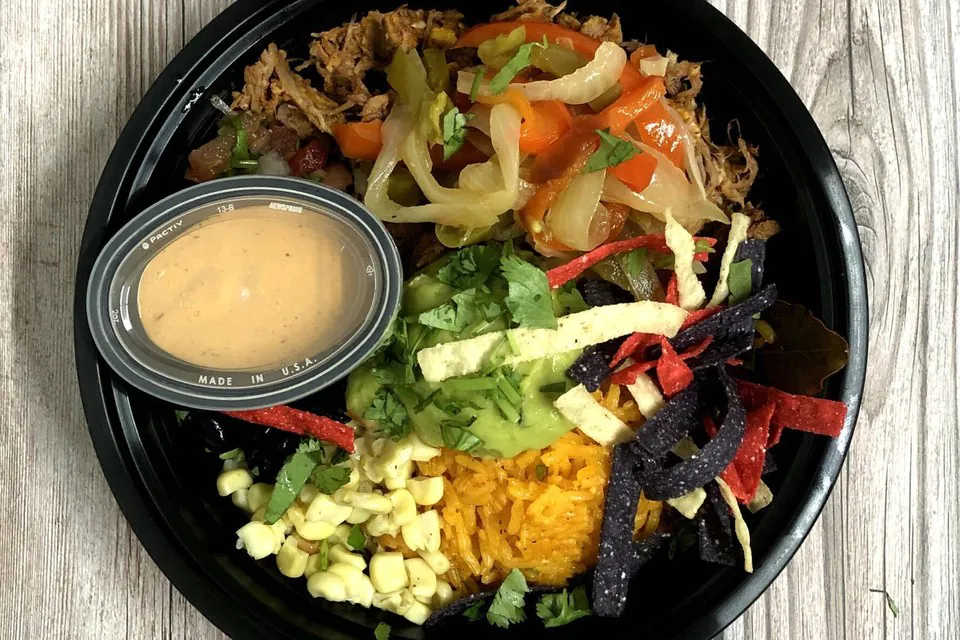 Overhead view of a black catering bowl with shredded pork carnitas, sauteed peppers and onions, Mexican rice, sweet corn, guacamole, and red and blue tortilla strips, with a small cup of chipotle crema on the side