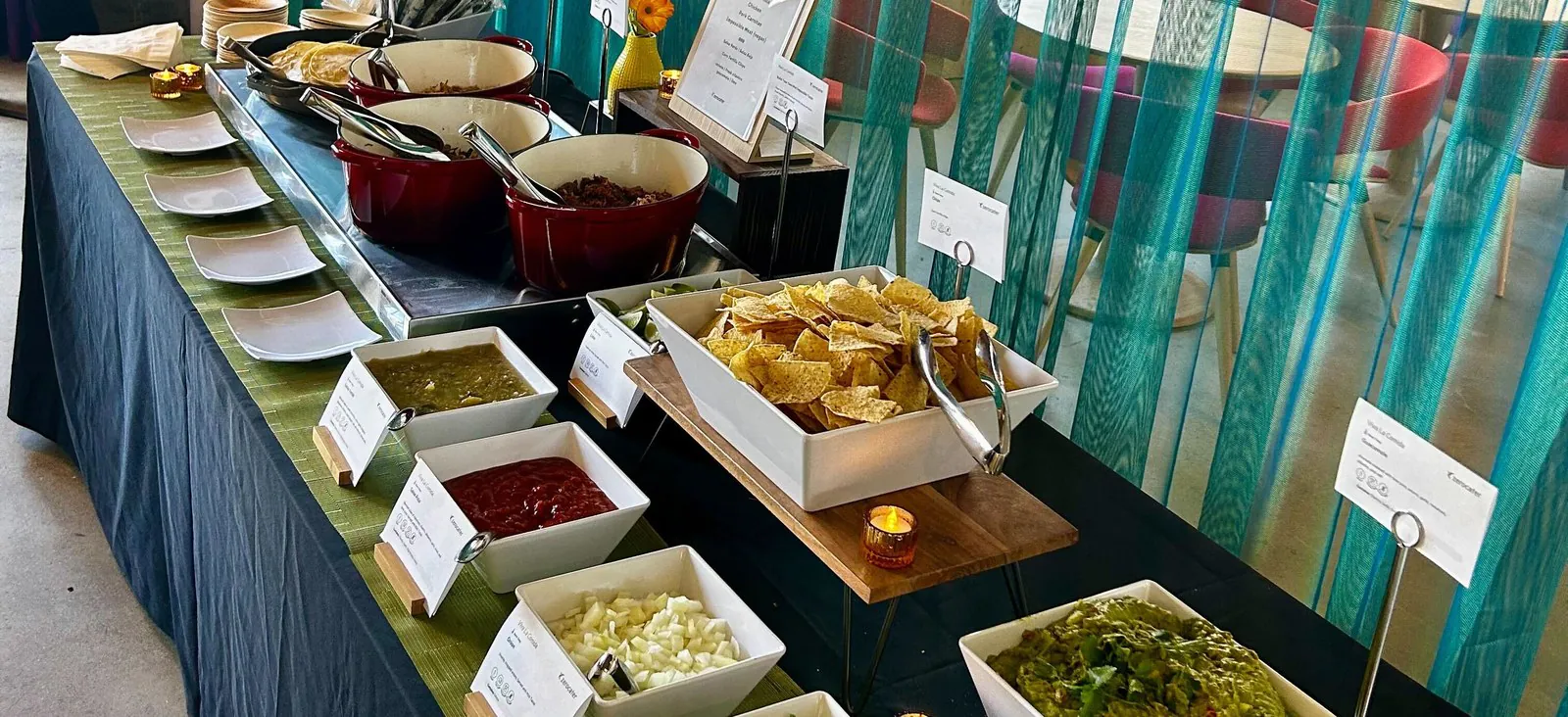 Editorial Tex-Mex office buffet line with tortilla chips on a wood riser, ramekins of salsa verde, salsa roja, white onion, fresh guacamole, and a chafing pot of refried beans on a long table with a green runner and teal curtains in the background