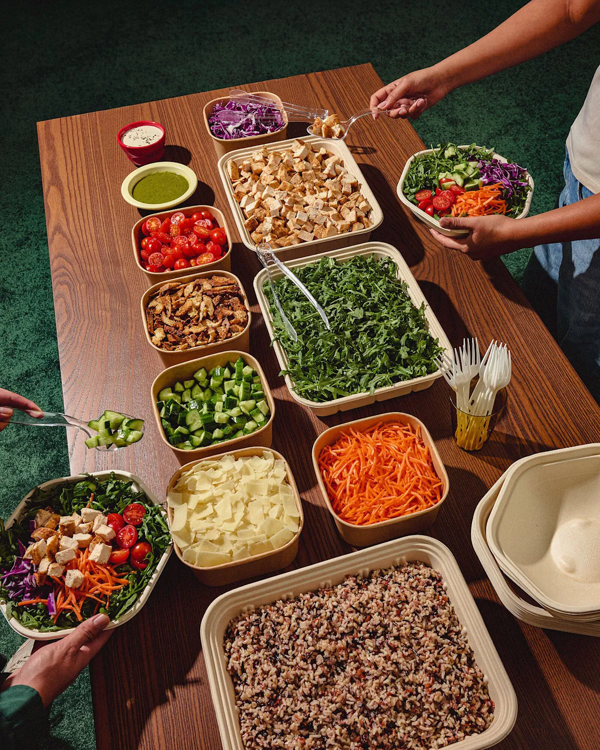 Sweetgreen catering buffet build line with compostable kraft bowls of cherry tomatoes, croutons, kale, cucumbers, carrots, mushrooms, red cabbage and quinoa arranged on a wood table