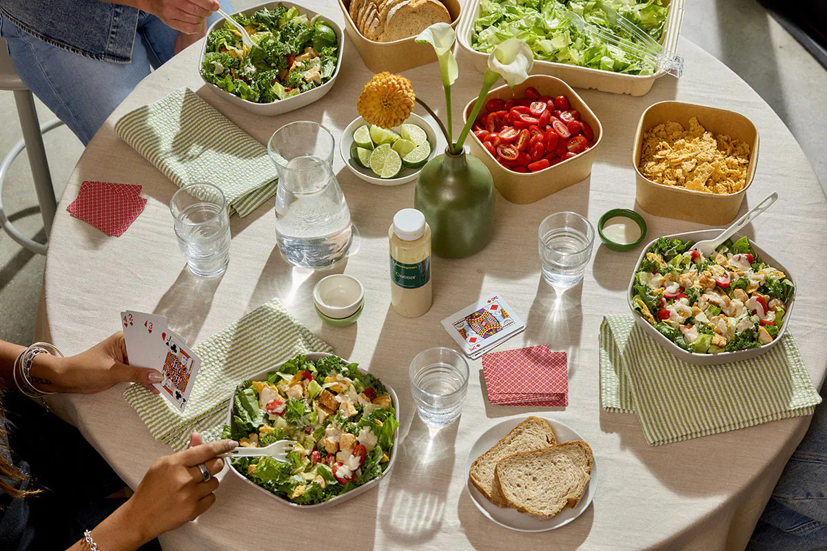 Sweetgreen catering at a meeting table with individually packaged warm bowls, a family-style platter of mixed greens, a glass water carafe, sliced bread, and ramekins of dressing arranged on a sage table runner