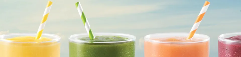 Row of four fresh-blended Tropical Smoothie Cafe smoothies in clear glass cups with paper straws: a yellow mango smoothie with a yellow striped straw, a deep green spinach detox smoothie with a green striped straw, an orange tropical smoothie with an orange striped straw, and a deep red berry smoothie, against a soft sky background
