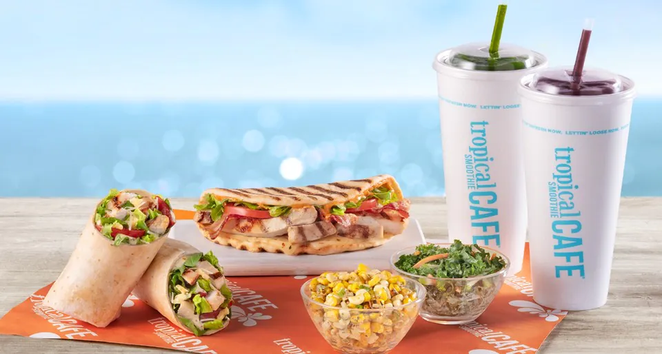 Tropical Smoothie Cafe catering spread on an orange tropical-CAFE branded mat with two chicken wraps, a grilled flatbread sandwich on a wood board, a small bowl of jalapeño corn slaw, a small bowl of kale and apple slaw, and two tall white Tropical Smoothie Cafe branded cups with green and red smoothies, beach background