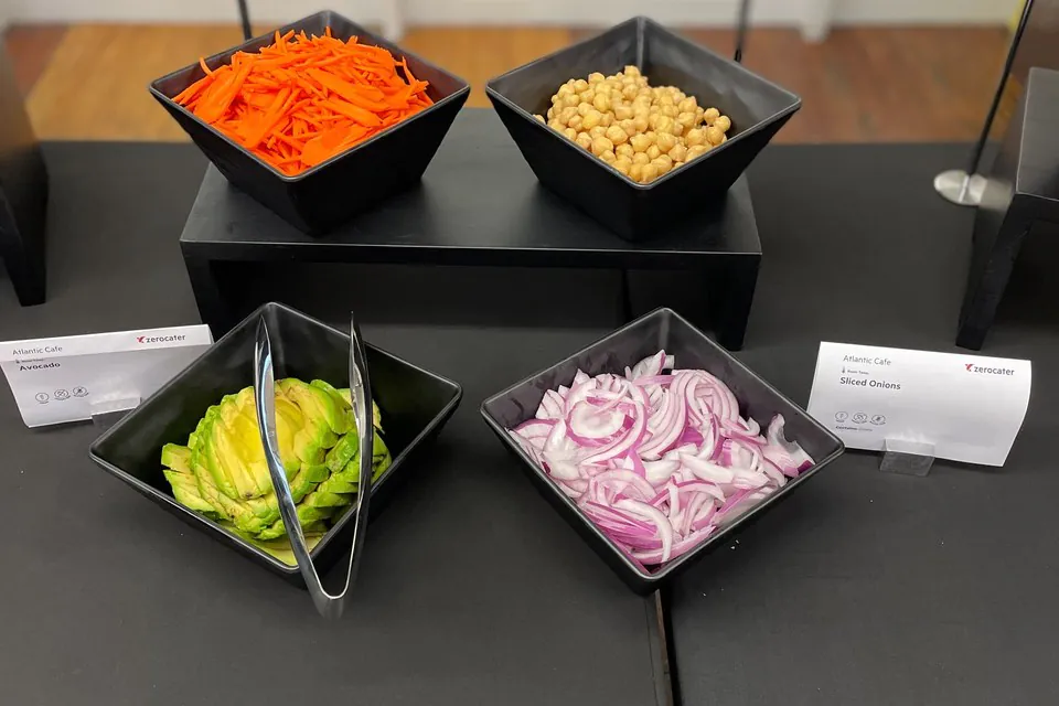 Build-your-own bowl bar with black square bowls of grilled tofu, chickpeas, avocado, and sliced red onions on a dark catering table with Zerocater menu cards