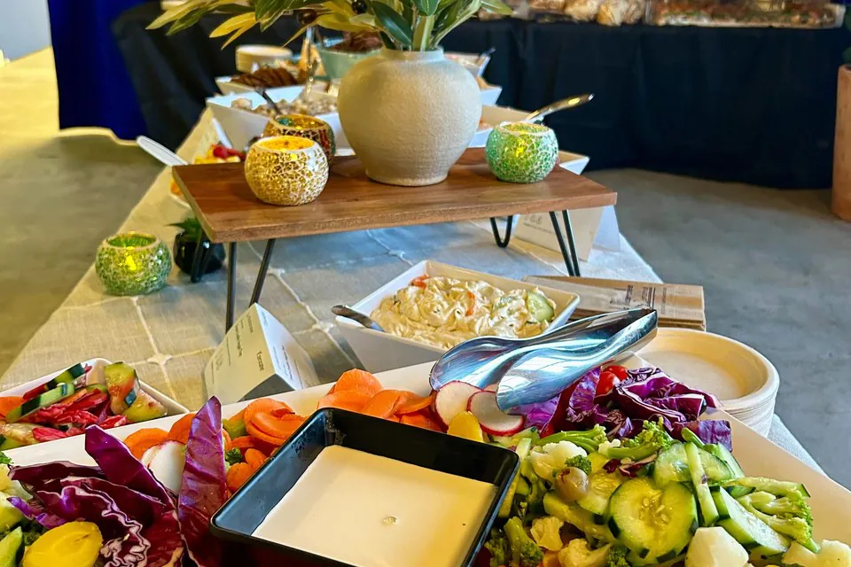 Crudite and hummus mezze platter with carrots, cucumber, red cabbage, radish, broccoli, and cauliflower around a white dipping sauce on a long catering table with olive branch centerpiece