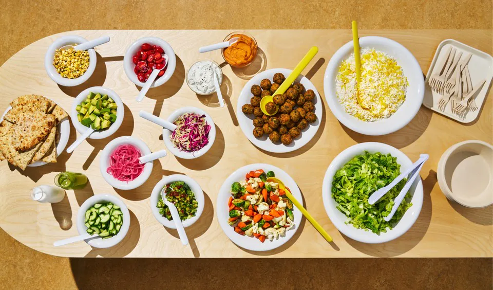 A CAVA Group Bowl Bar catering setup on a light wood table with bowls of falafel, fire-roasted corn, tomatoes, cucumbers, slaw, feta, dips and chopped greens with serving spoons
