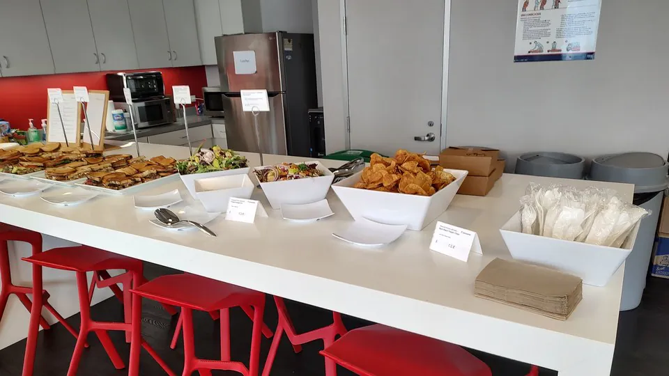 Office buffet table with platters of sliced deli sandwiches, fresh salads in white bowls, and a bowl of chips with plates and utensils