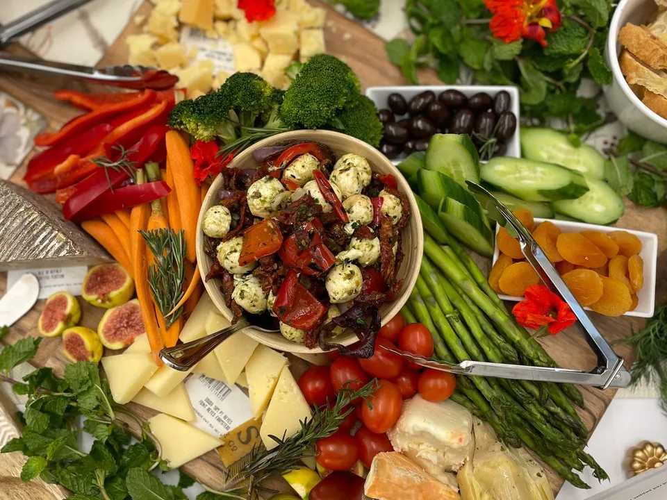Overhead view of an abundant grazing board with marinated mozzarella, aged and soft cheeses, crudite, asparagus, cherry tomatoes, olives, dried apricots, figs, and edible flowers on a wooden board