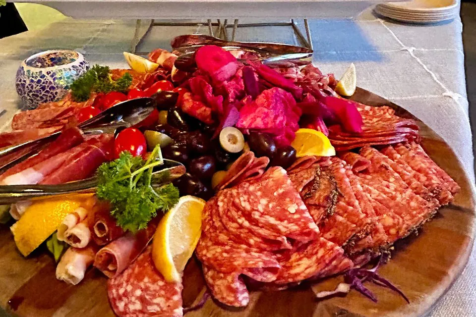 Close-up of a round charcuterie board with fanned salami, prosciutto rolls, soppressata, olives, pickled onions, lemon wedges, and parsley with serving tongs