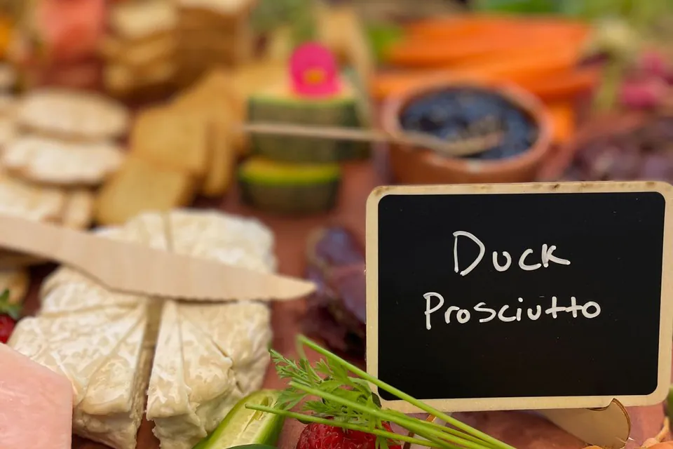 A close-up of a hand-lettered chalkboard label reading Duck Prosciutto on a charcuterie station next to brie, crackers, carrots, and cucumber